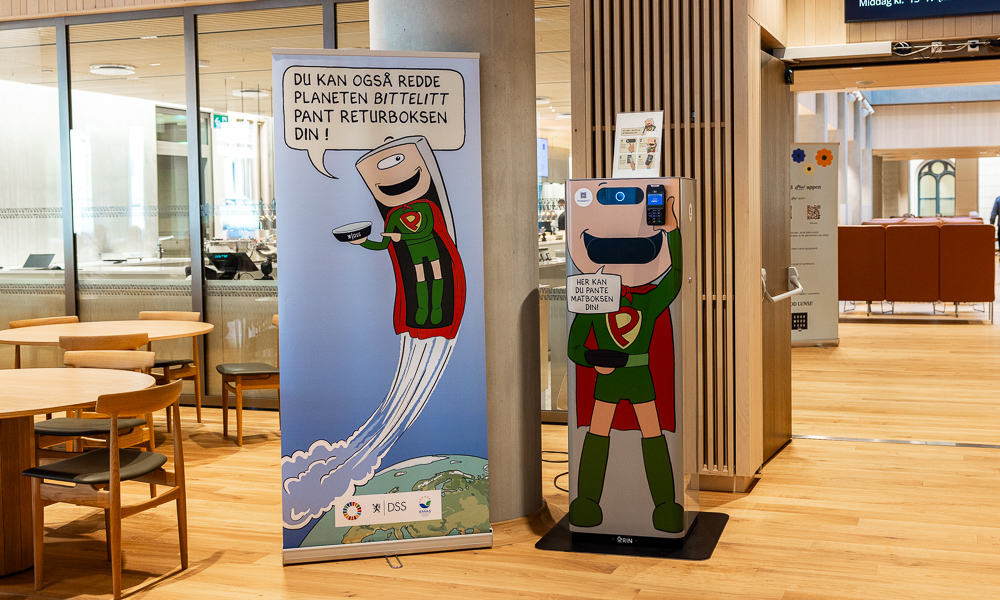 En panteautomat for gjenbruksemballasje ved siden av en stor plakat. På automaten står det skrevet "Her kan du pante matboksen din". På plakaten ser man karakteren "pantus" i superheltdrakt med tilhørende tekst som leser "Du kan også redde planeten bittelitt pant returboksen din!".