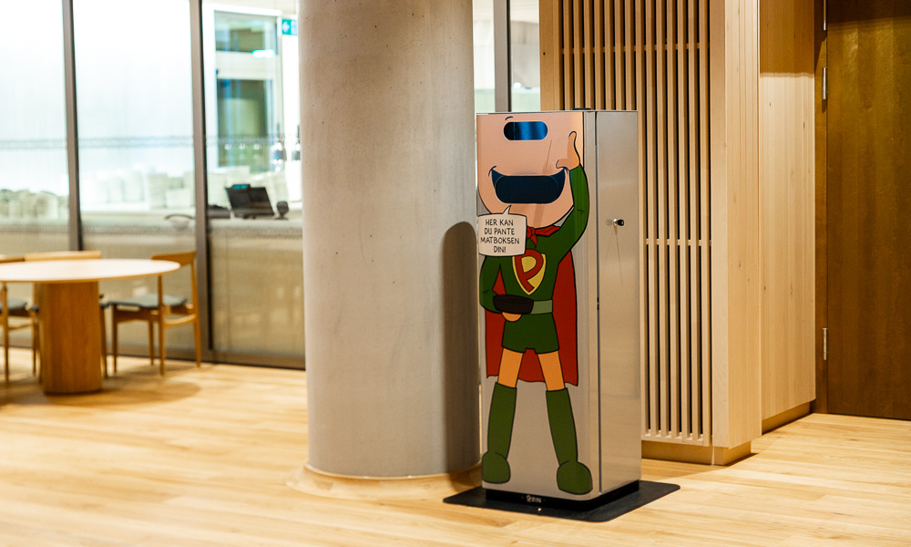 En panteautomat for gjenbruksemballasje ved siden av en stor pillar. Plasser rett ved kantinen tilhørende nytt regjeringskvartal. På automaten står det skrevet "Her kan du pante matboksen din".