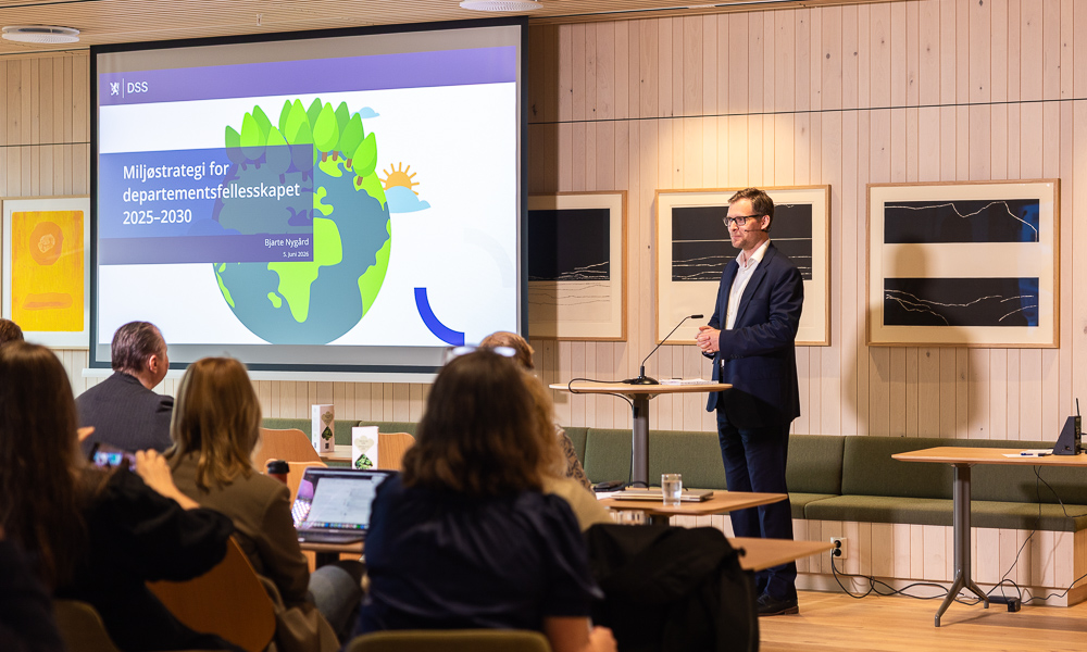 En ansatt står foran et publikum og presenterer. På skjermen ser man følgende tekst "Miljøstrategi for departementsfelleskapet 2025-2030.
