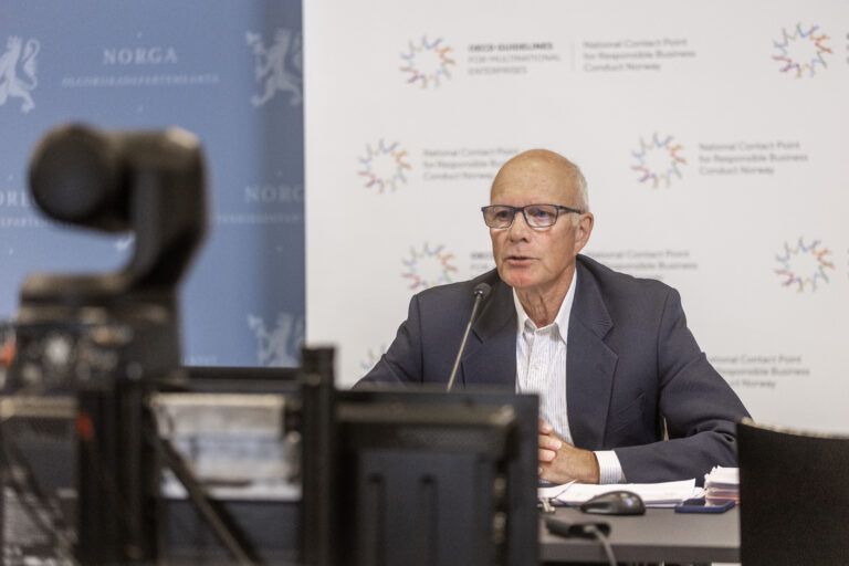 Mann i dress sitter foran skjermer og snakker til et kamera i forgrunnen.