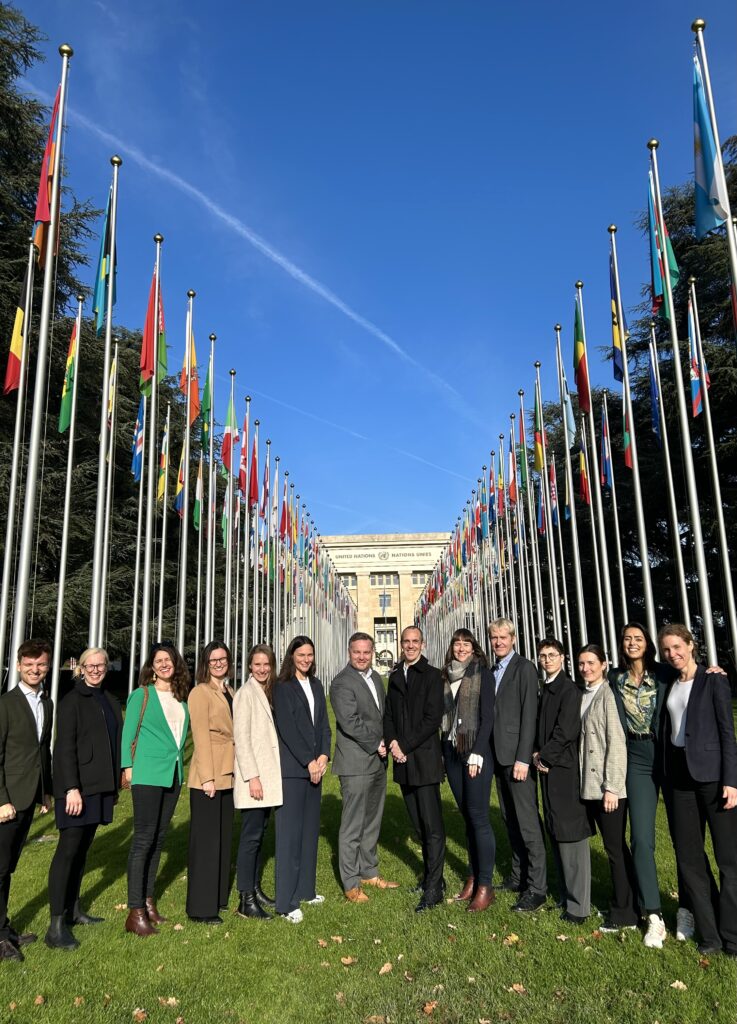 En rekke med mennesker foran alle FNs flagg i Genève. Sol og blå himmel.