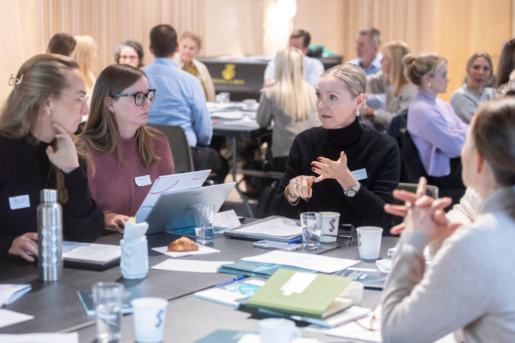 Tre personer retter ser oppmerksomt mot en fjerde under ivrig samtale rundt et bord under workshopen