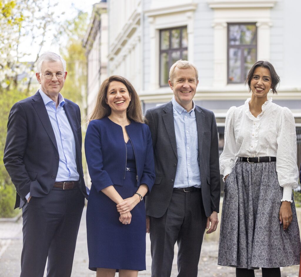 Fire personer på rekke utendørs foran bygård. To menn og to damer.