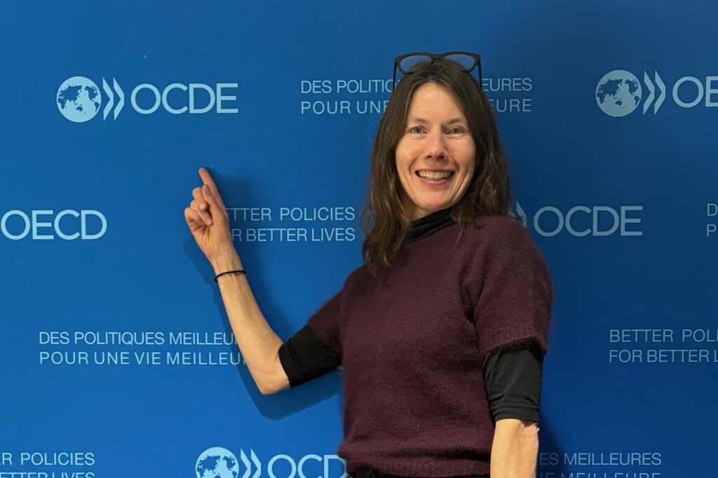 Bilde av dame med briller foran blå fotovegg med OECD i tekst.